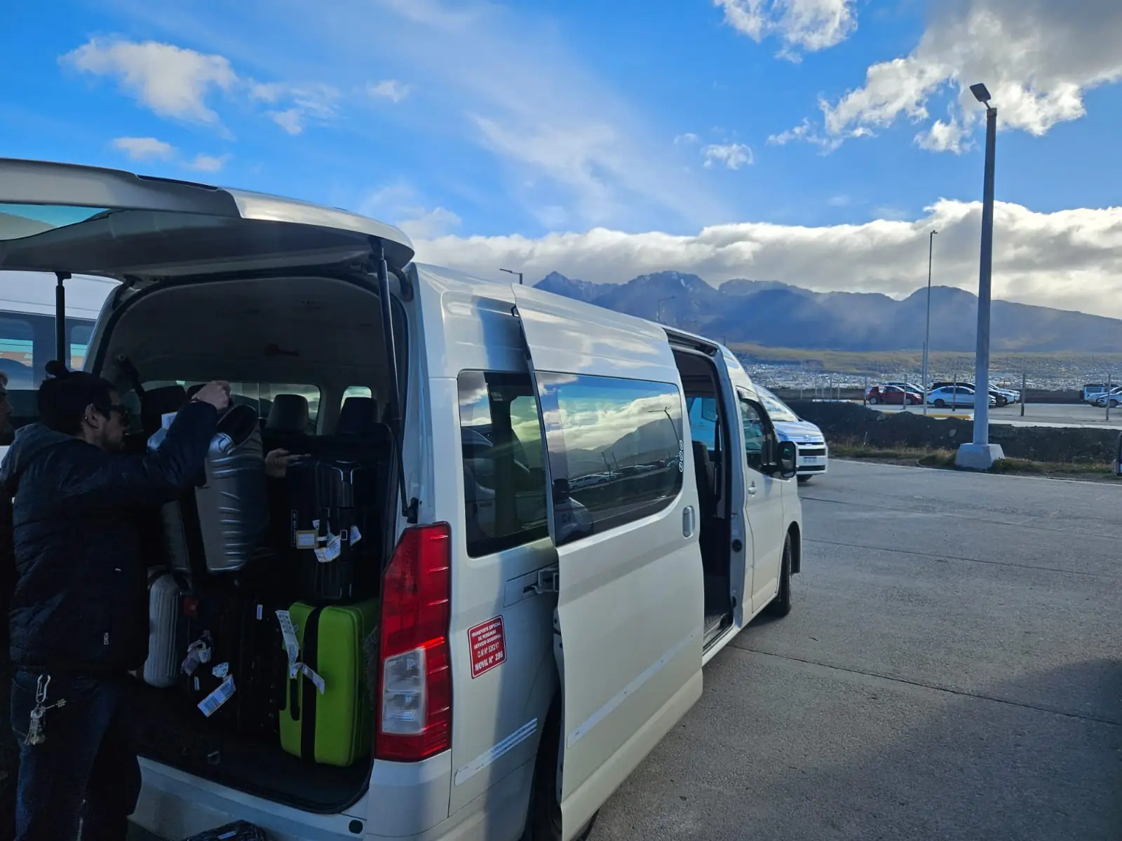 Driver loading suitcases into the private transfer van at the Ushuaia port parking, with Fuegian mountains behind